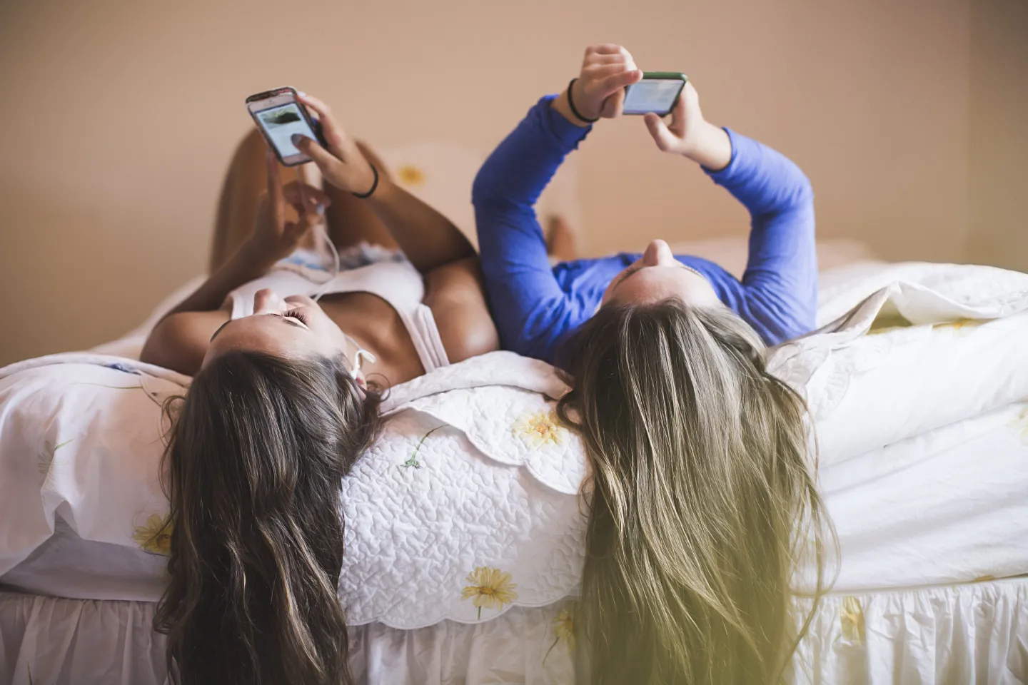 Two young women lying side by side, hair flowing down the edge of the bed in the foreground, both holding smartphones