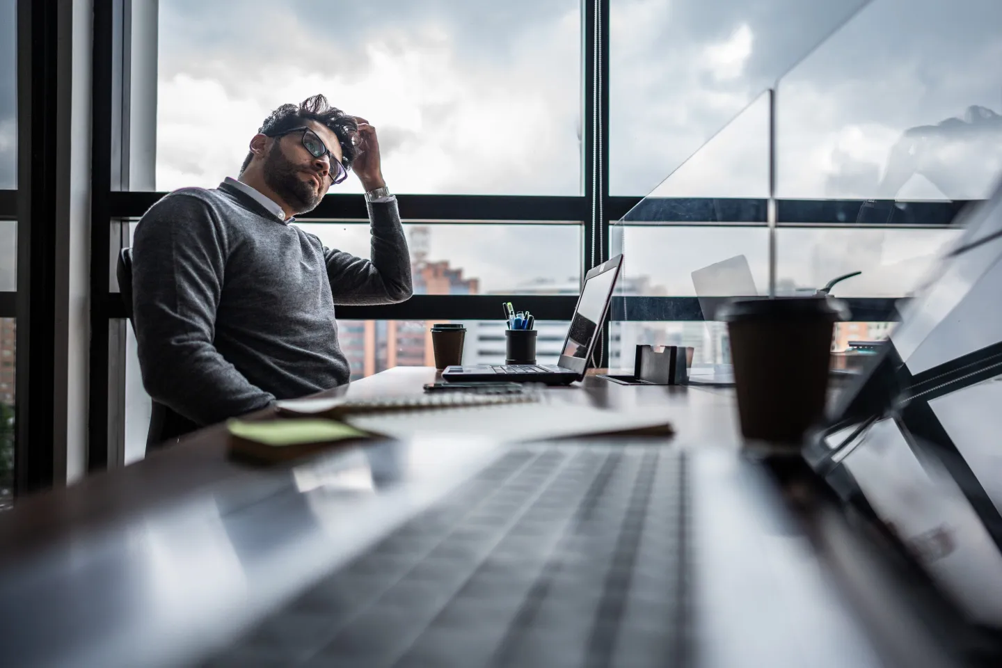 Man at desk in front of large window with hand in hair, looking distracted