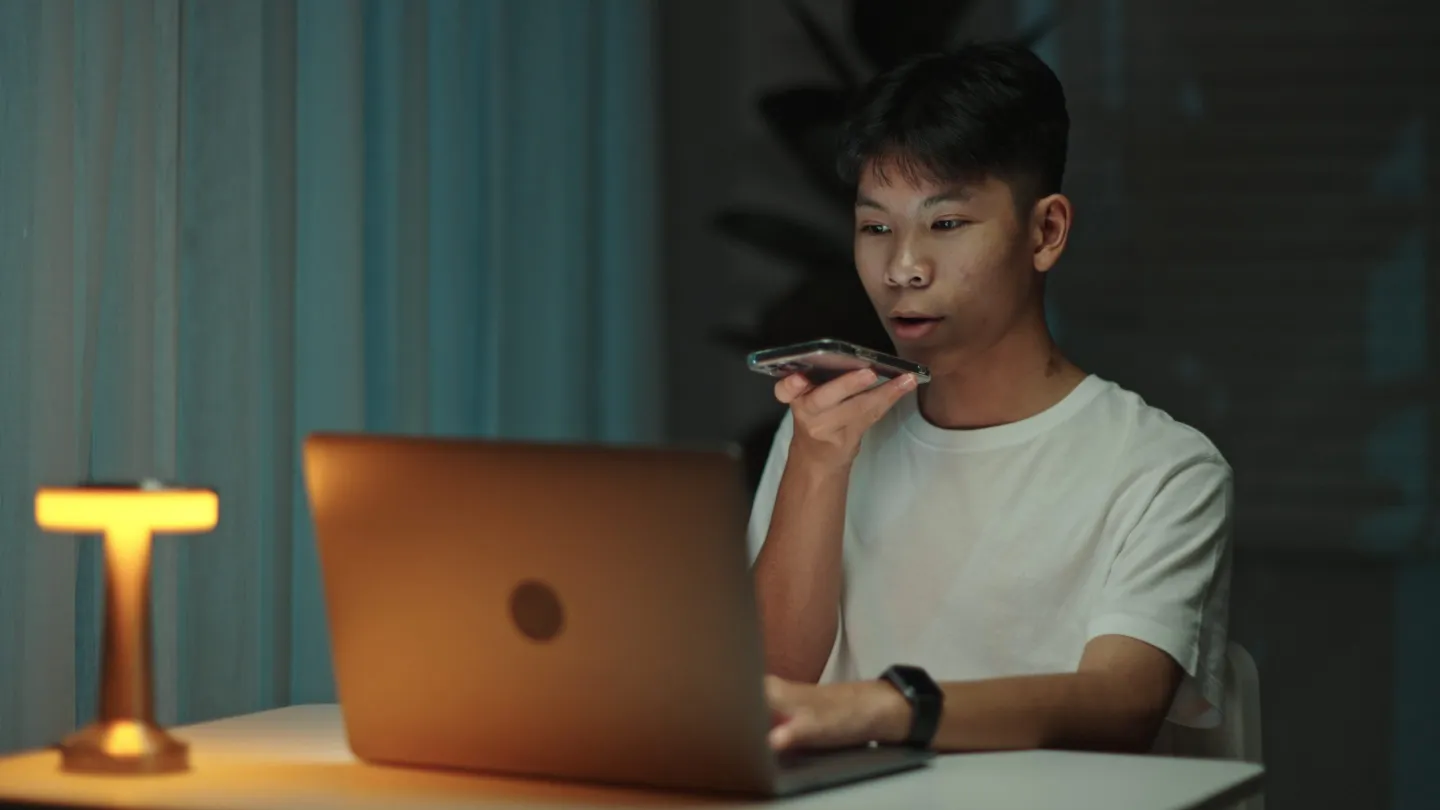 A teenage boy sitting at a desk with a laptop open in front of him and talking into his phone.