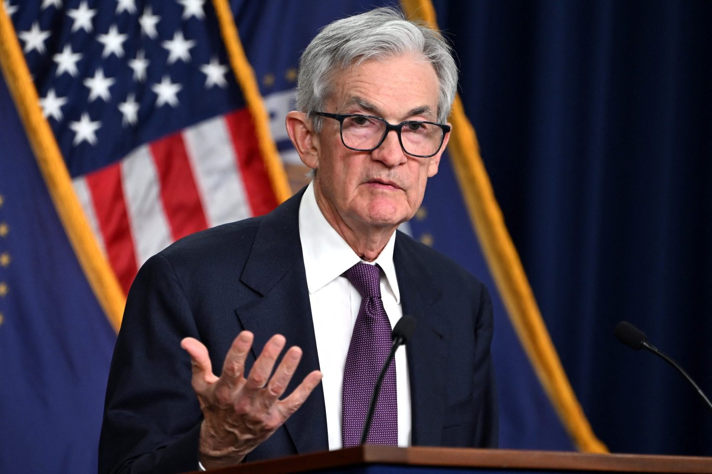 Jerome Powell raises his left hand as he speaks at a press conference.