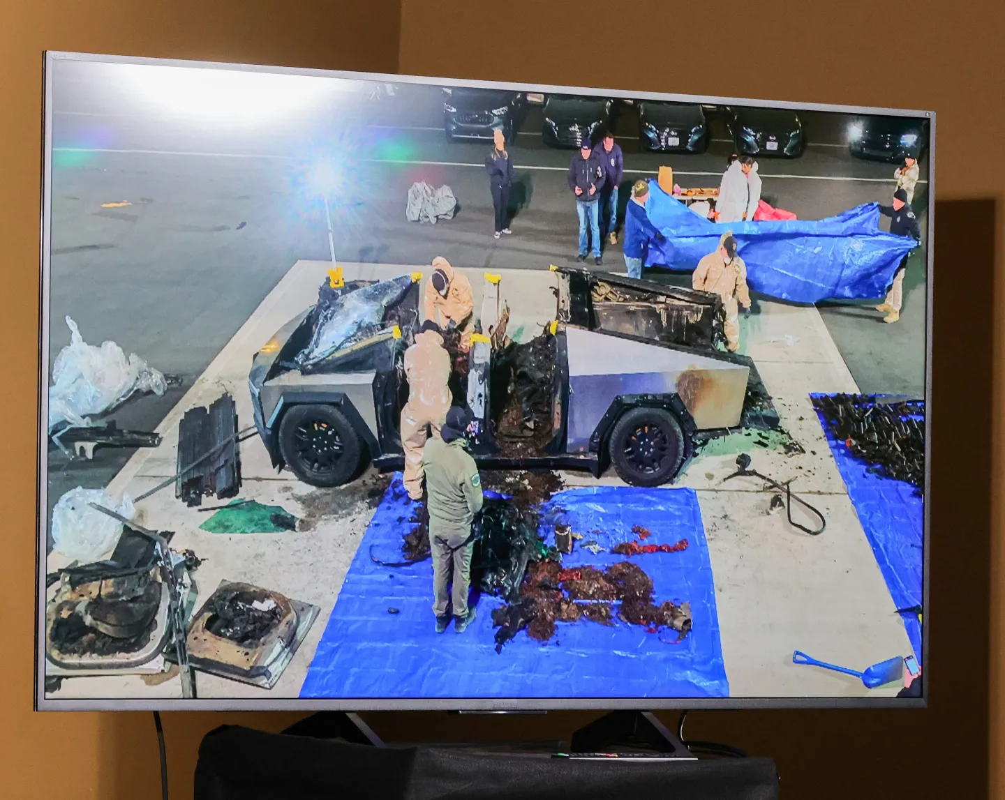 Photo of a TV screen showing law enforcement forensic experts searching the remains of a burnt out Tesla Cybertruck for evidence.