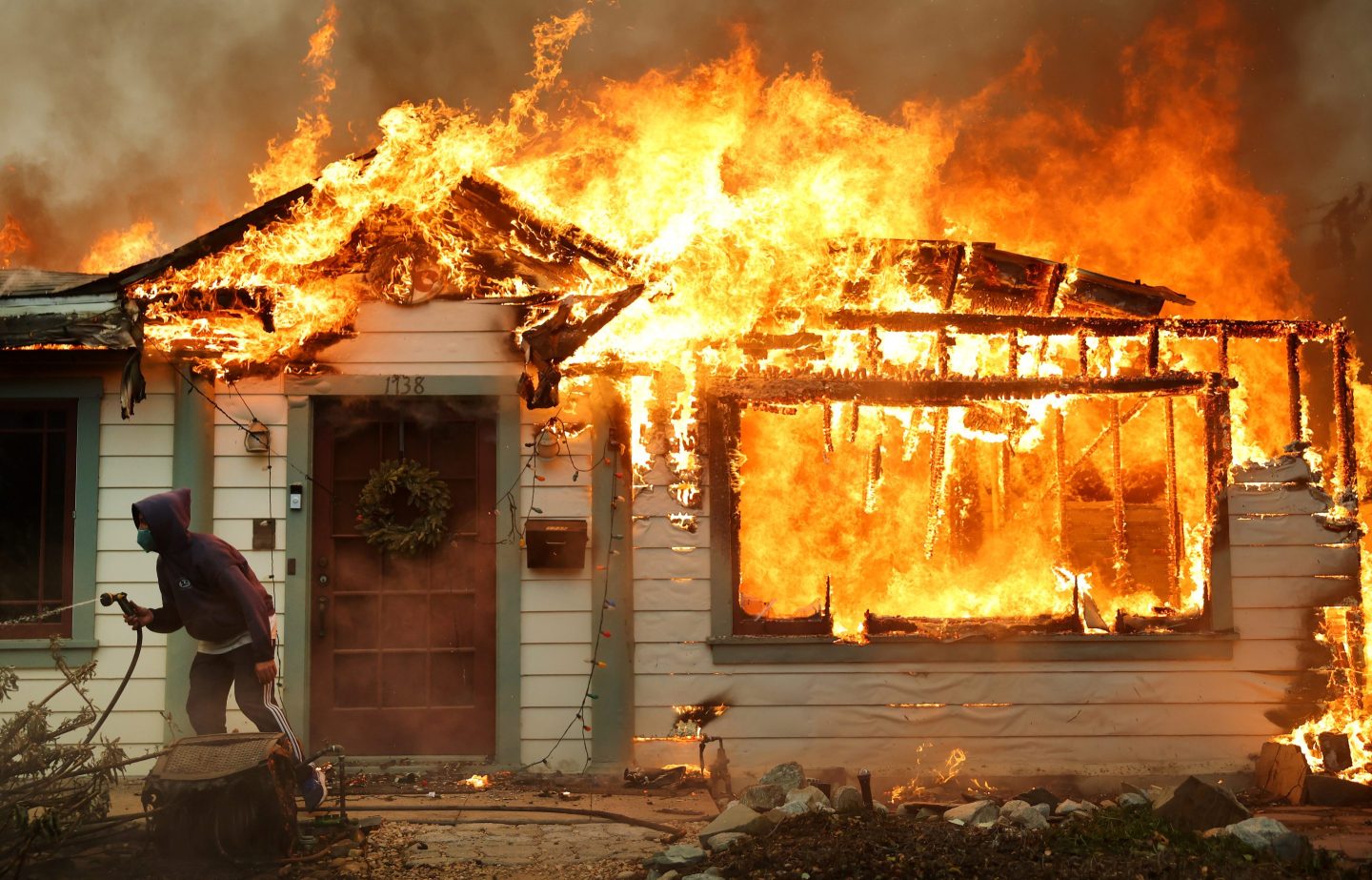 A person stands outside a burning home, holding a garden hose