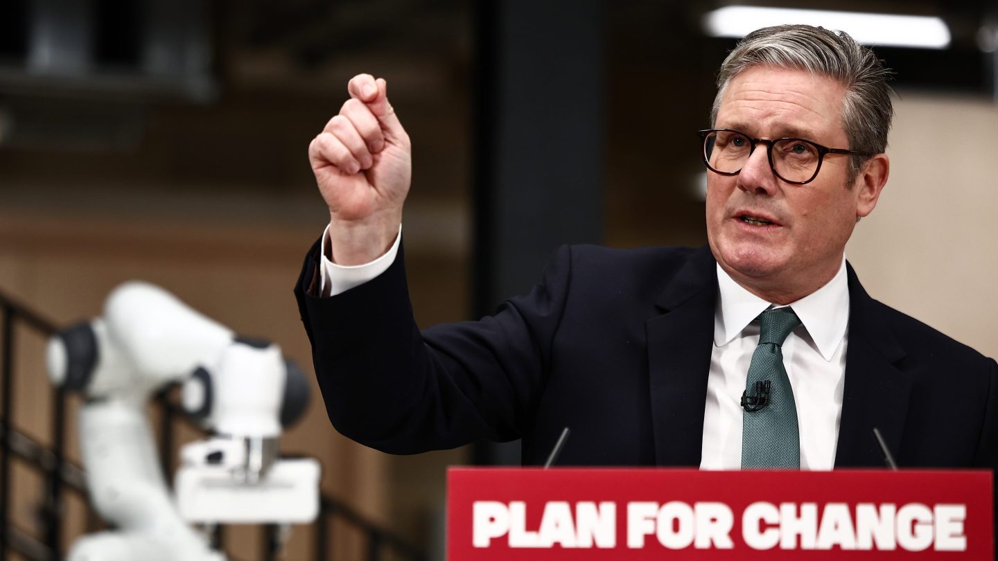 U.K. Prime Minister Keir Starmer speaking before a podium with a sign saying &quot;Plan for Change&quot; on it and holding his right arm up in a gesture, while a robot arm is visible in the background.