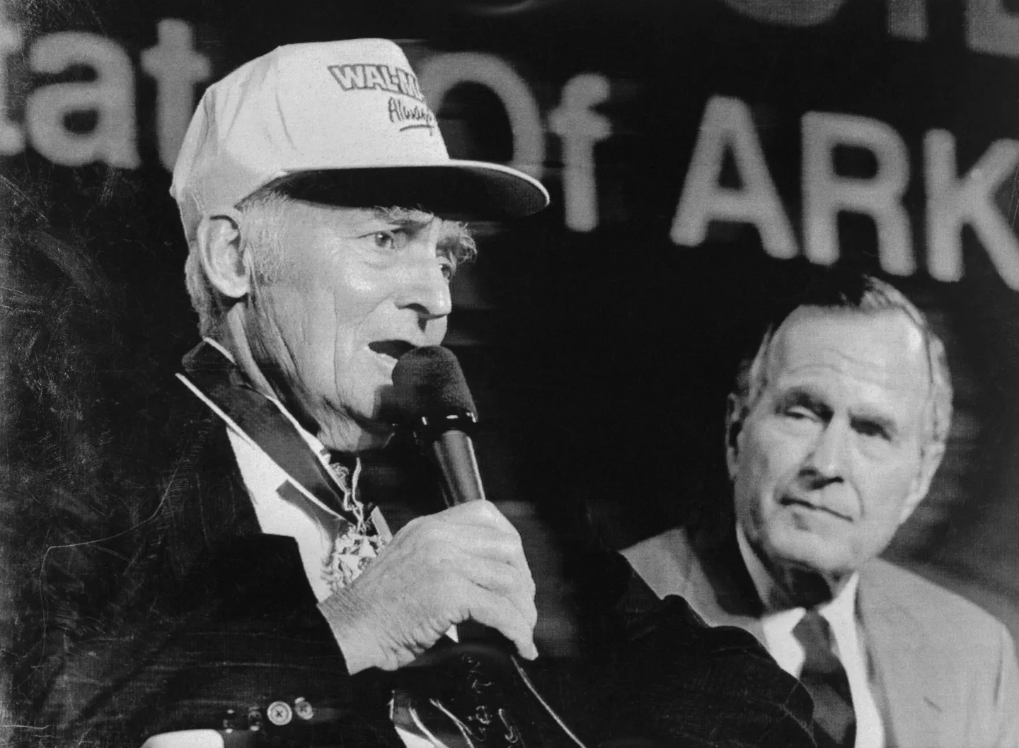 Sam Walton in his iconic trucker hat speaks into a mic as President George H.W. Bush looks on.