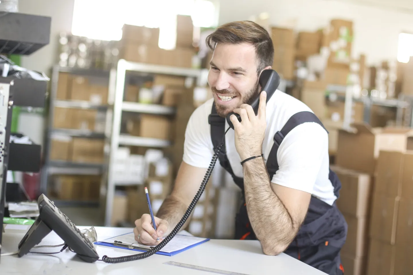 Smiling Caucasian male worker working in warehouse, talking on a landline phone and making a list.