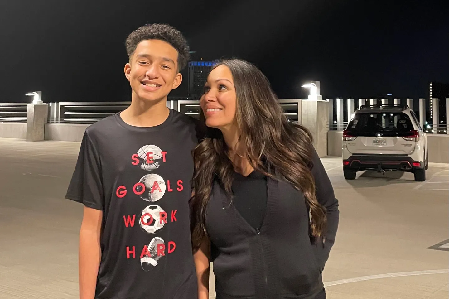 Tall young man posing with a shorter woman, his mom, outside. Both are smiling.