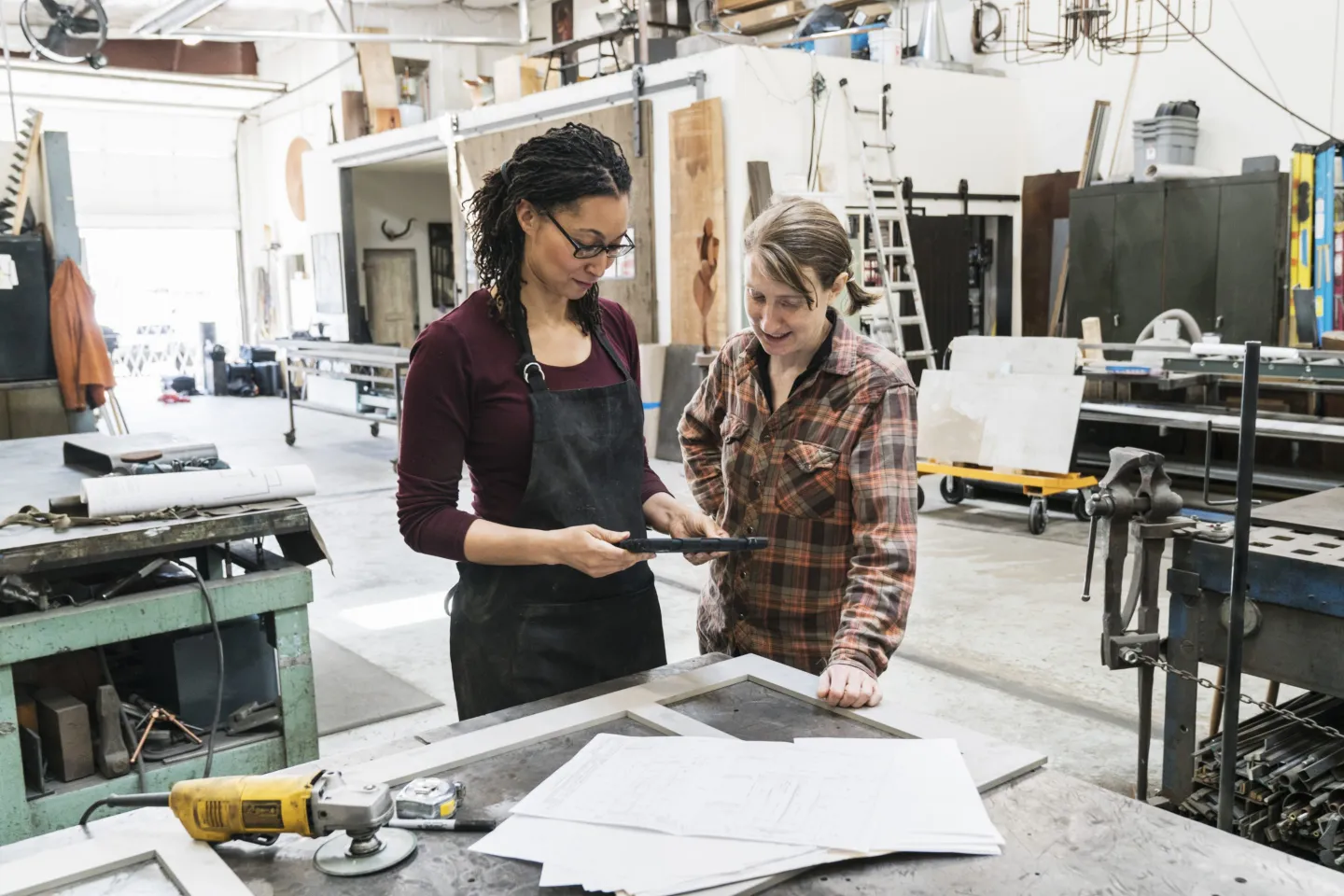 Two women standing at workbench in metal workshop, holding digital tablet.