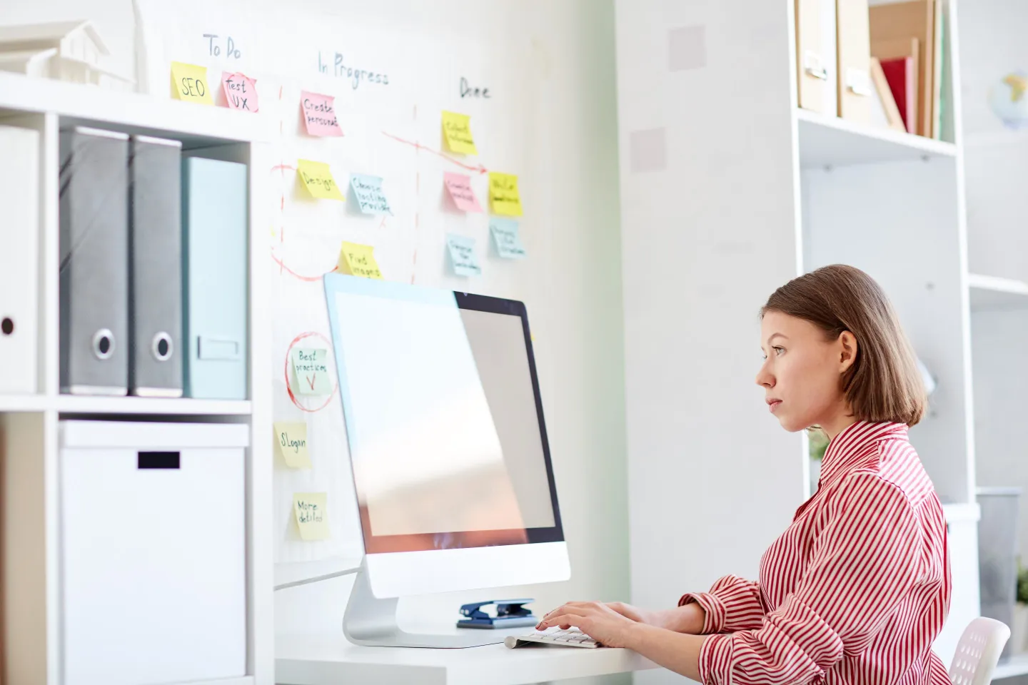 Side view of young Caucasian woman in striped shirt sitting at desk and working on computer in modern office