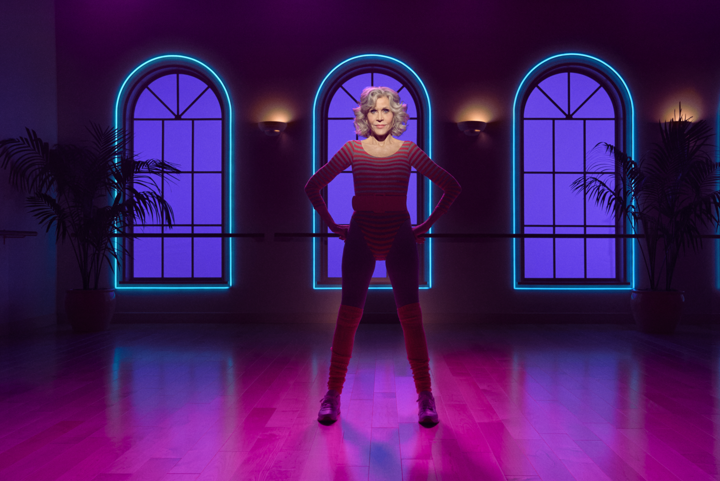 Jane Fonda standing in a purple-lit studio with her hands on her hips